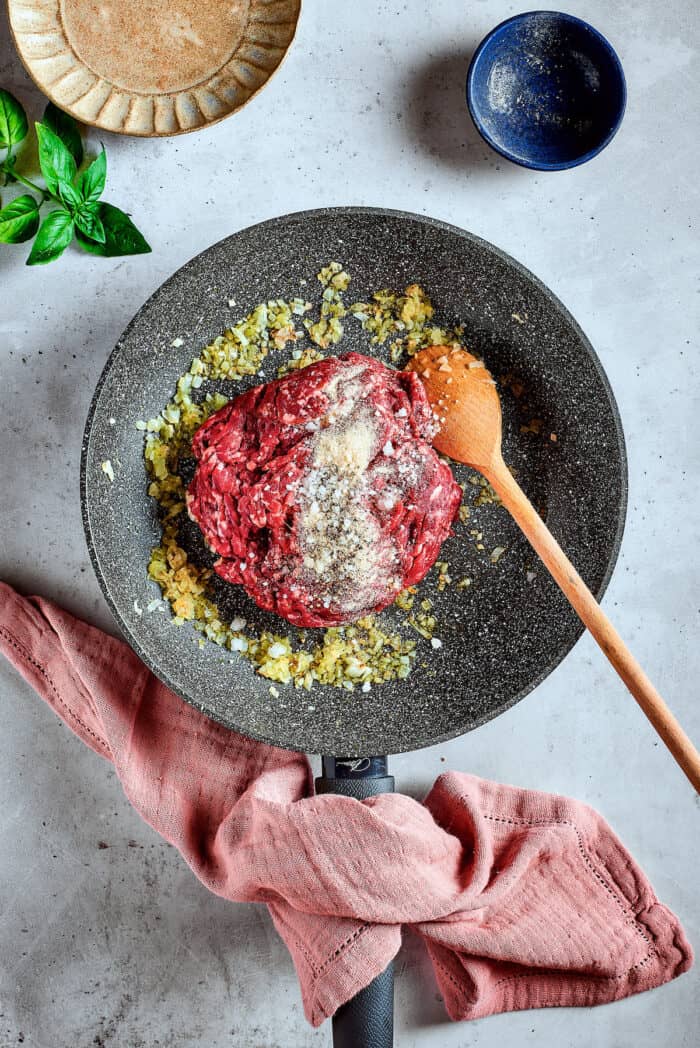 Cozy Stuffed Shells with Meat are packed with plenty of ground beef and a creamy, pesto-spiked filling to make a hearty meal. Meat is placed in a skillet with spices with a wooden spoon.