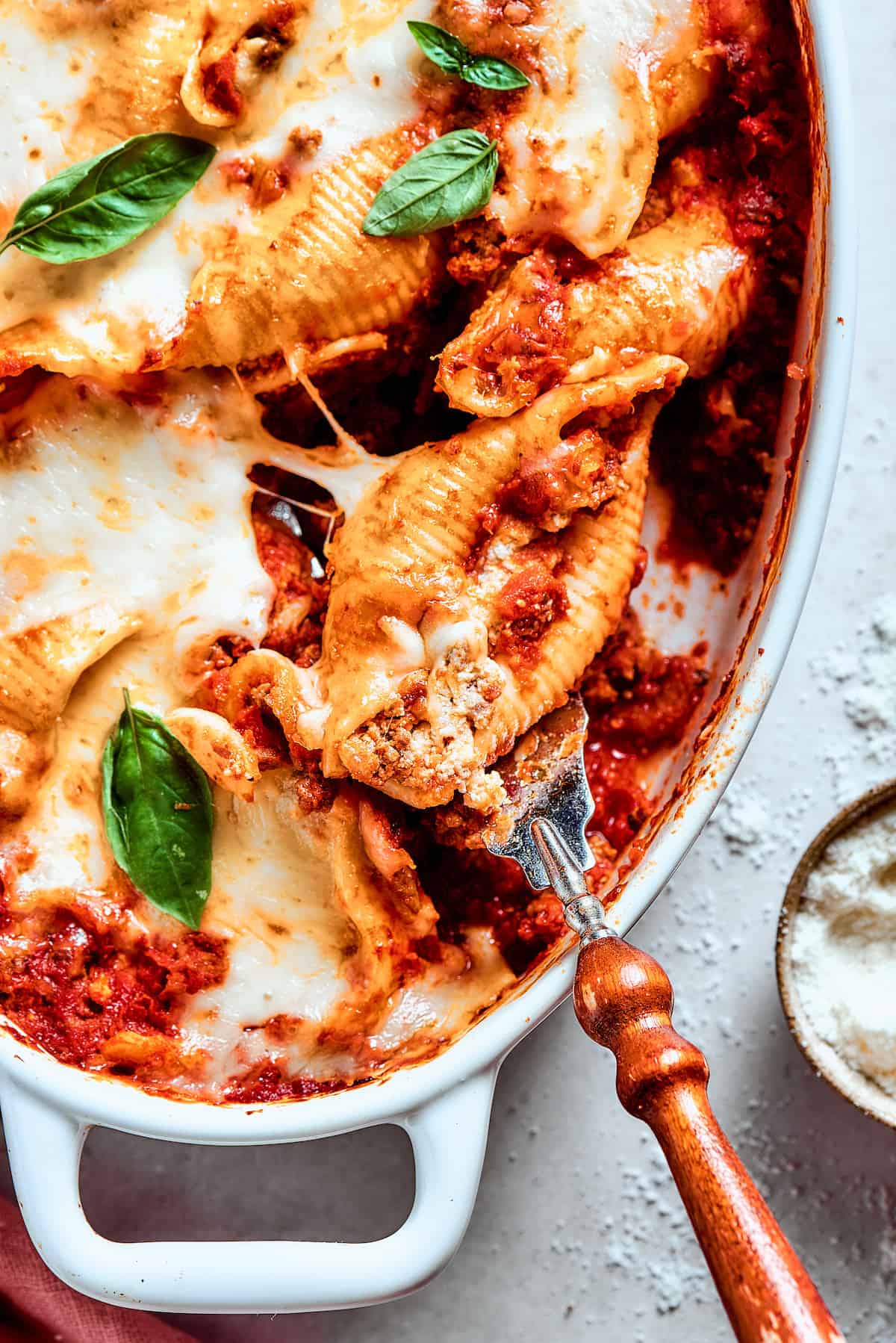 Cozy Stuffed Shells with Meat are packed with plenty of ground beef and a creamy, pesto-spiked filling to make a hearty meal. A serving utensil lifts out a stuffed shell with meat from the pan.