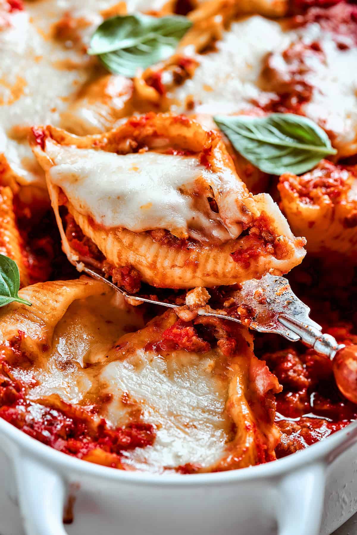 Cozy Stuffed Shells with Meat are packed with plenty of ground beef and a creamy, pesto-spiked filling to make a hearty meal. A serving utensil lifts out a stuffed shell with meat from the pan.