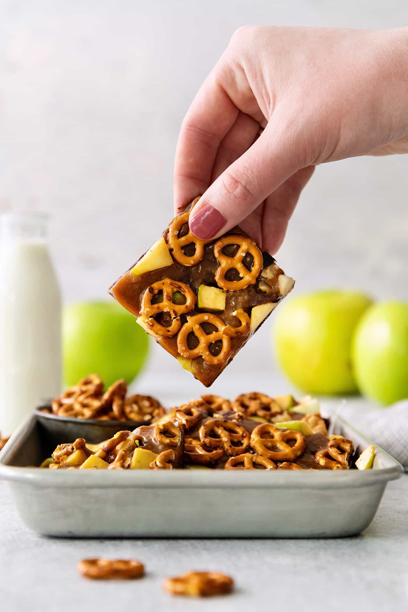 This Caramel Apple Bark is a fun treat to make when you want the flavor of a fresh caramel apple without the hassle. A hand lifts up a piece of caramel apple bark from a try with more pieces of bark with green apples in the background.