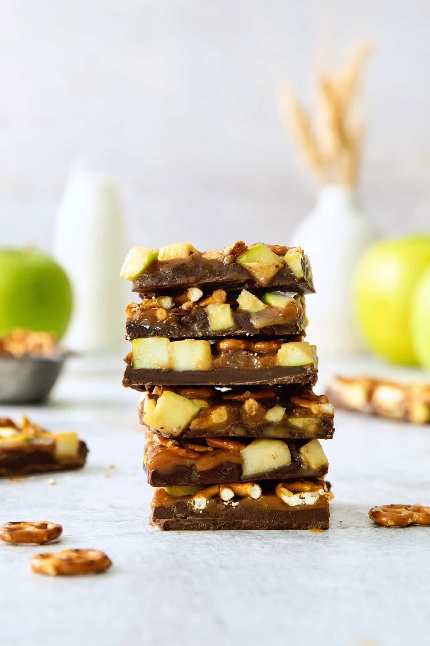 This Caramel Apple Bark is a fun treat to make when you want the flavor of a fresh caramel apple without the hassle. A stack of caramel apple bark with pretzels with apples in the background.