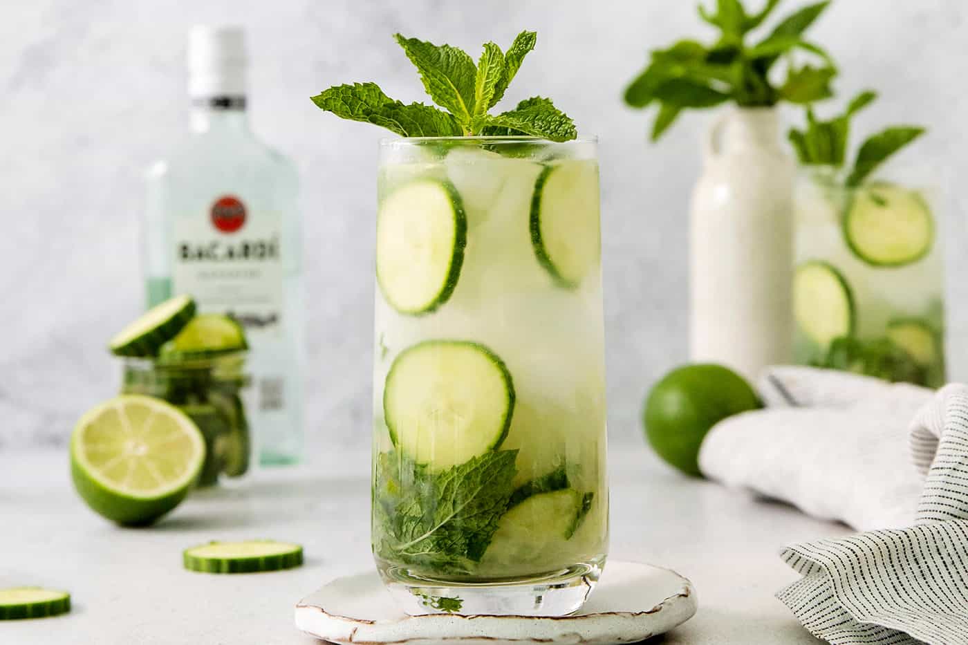 A Cucumber Mojito takes your classic mojito recipe and adds orange liqueur and cucumber slices for a refreshing twist on a minty favorite. Glasses of cucumber mojito topped with basil are shown with a bottle of rum in the background.