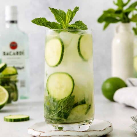Glasses of cucumber mojito topped with basil are shown with a bottle of rum in the background.