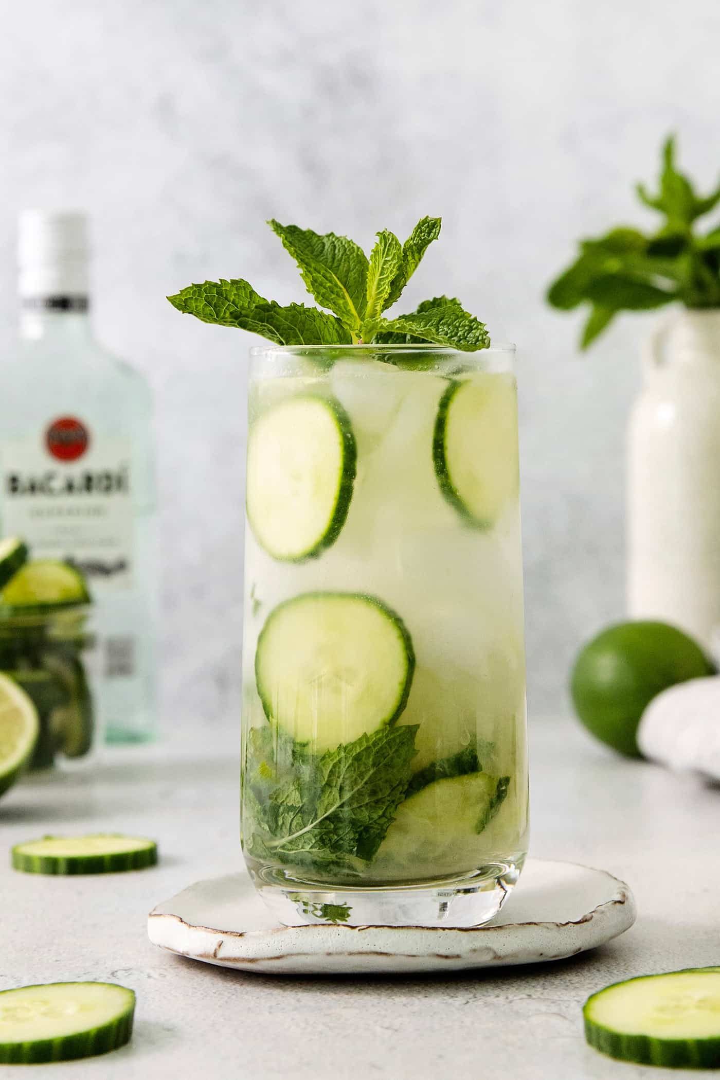 A Cucumber Mojito takes your classic mojito recipe and adds orange liqueur and cucumber slices for a refreshing twist on a minty favorite. Glasses of cucumber mojito topped with basil are shown with a bottle of rum in the background.