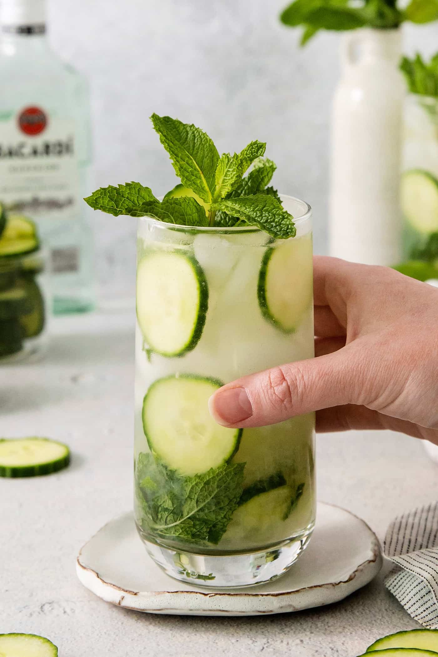A Cucumber Mojito takes your classic mojito recipe and adds orange liqueur and cucumber slices for a refreshing twist on a minty favorite. A hand picks up a glass of cucumber mojito.