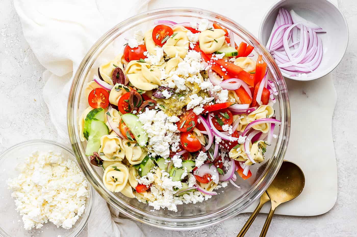 reek Tortellini Salad is full of fresh Greek flavors like lemon, olive oil, herbs, and feta cheese! This cold salad is great at picnics. A bowl of Greek tortellini salad is shown on a white table with serving spoons.