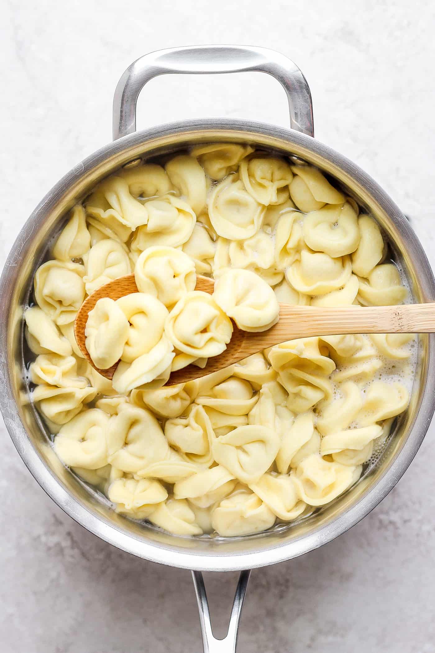 reek Tortellini Salad is full of fresh Greek flavors like lemon, olive oil, herbs, and feta cheese! This cold salad is great at picnics. A wooden spoon lifts tortellini out of a pot.