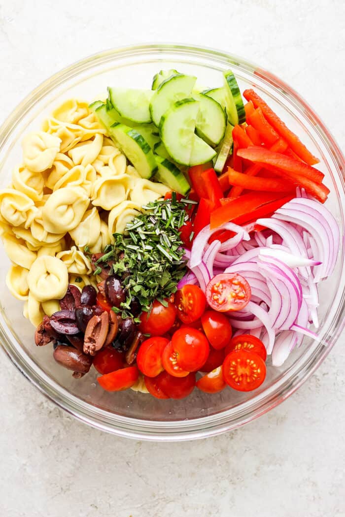 reek Tortellini Salad is full of fresh Greek flavors like lemon, olive oil, herbs, and feta cheese! This cold salad is great at picnics. Ingredients for Greek tortellini salad are shown in a glass bowl.