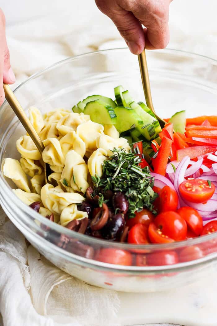 reek Tortellini Salad is full of fresh Greek flavors like lemon, olive oil, herbs, and feta cheese! This cold salad is great at picnics. Spoons mix together tortellini salad in a bowl.