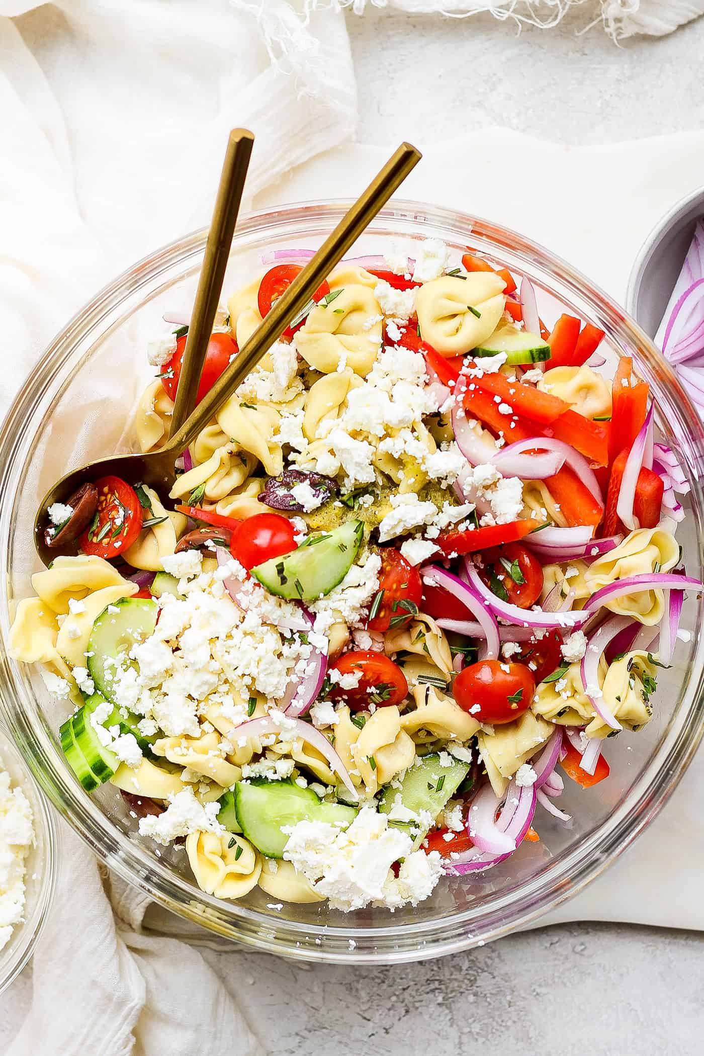 reek Tortellini Salad is full of fresh Greek flavors like lemon, olive oil, herbs, and feta cheese! This cold salad is great at picnics. A bowl of Greek tortellini salad with serving spoons.