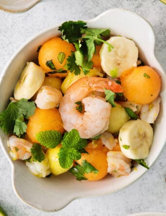 An overhead shot of a colorful bowl of tropical shrimp salad with fruit, shrimp, and mint leaves.