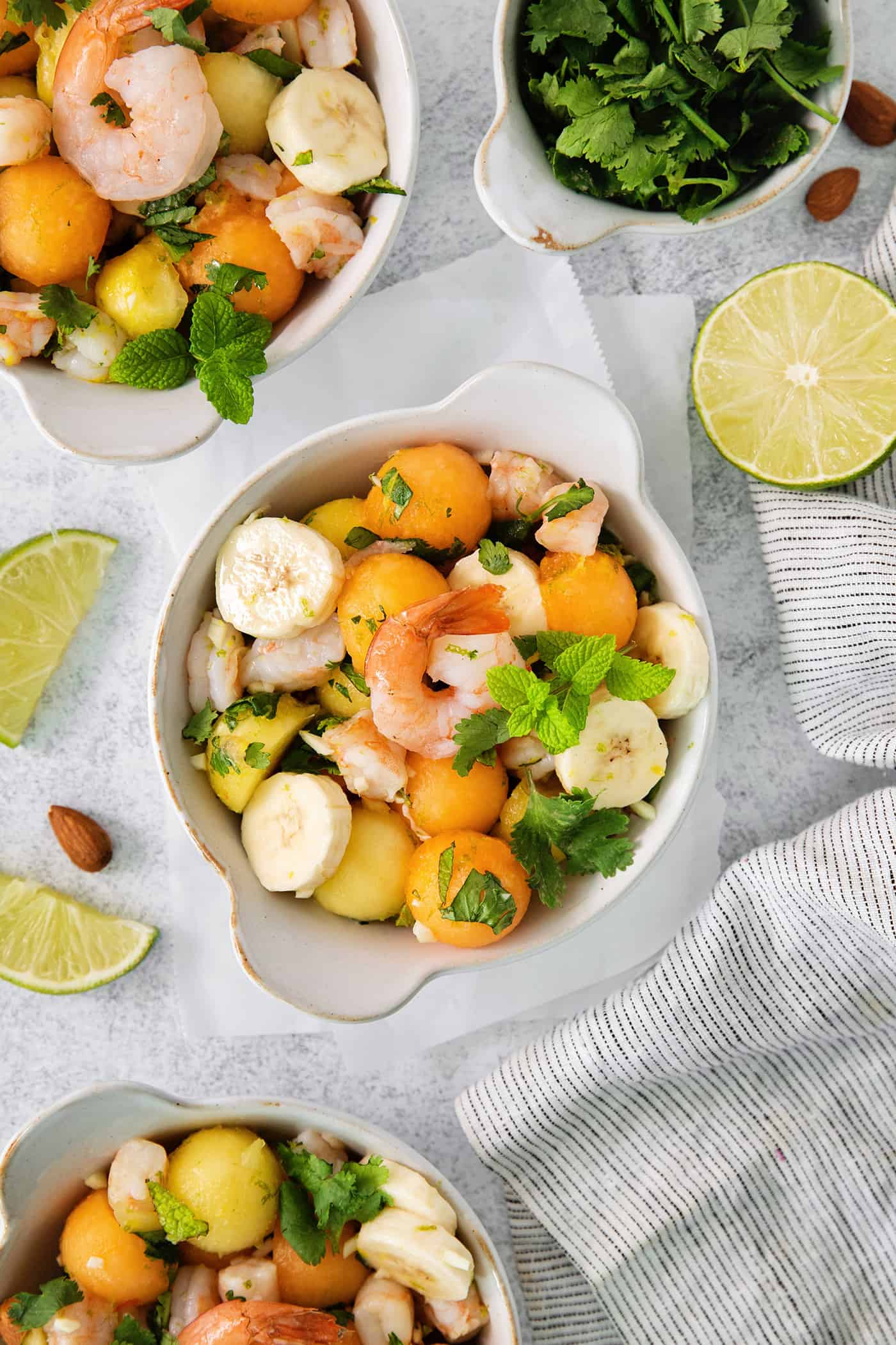 This Tropical Shrimp Salad has fresh, bright color and flavor. It's a unique, refreshing appetizer or salad recipe that's great for entertaining! An overhead shot of colorful bowls of tropical shrimp salad with fruit, shrimp, and mint leaves and limes on the side.