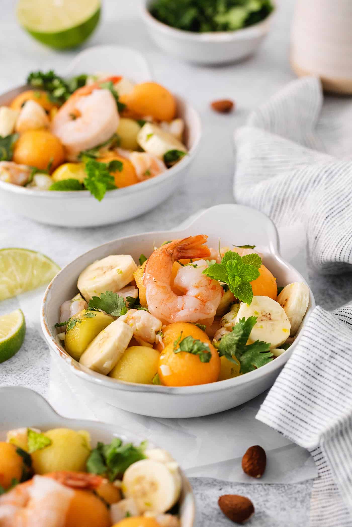 This Tropical Shrimp Salad has fresh, bright color and flavor. It's a unique, refreshing appetizer or salad recipe that's great for entertaining! Colorful bowls of tropical shrimp salad with fruit, shrimp, and mint leaves and limes on the side are shown on a table with napkins.