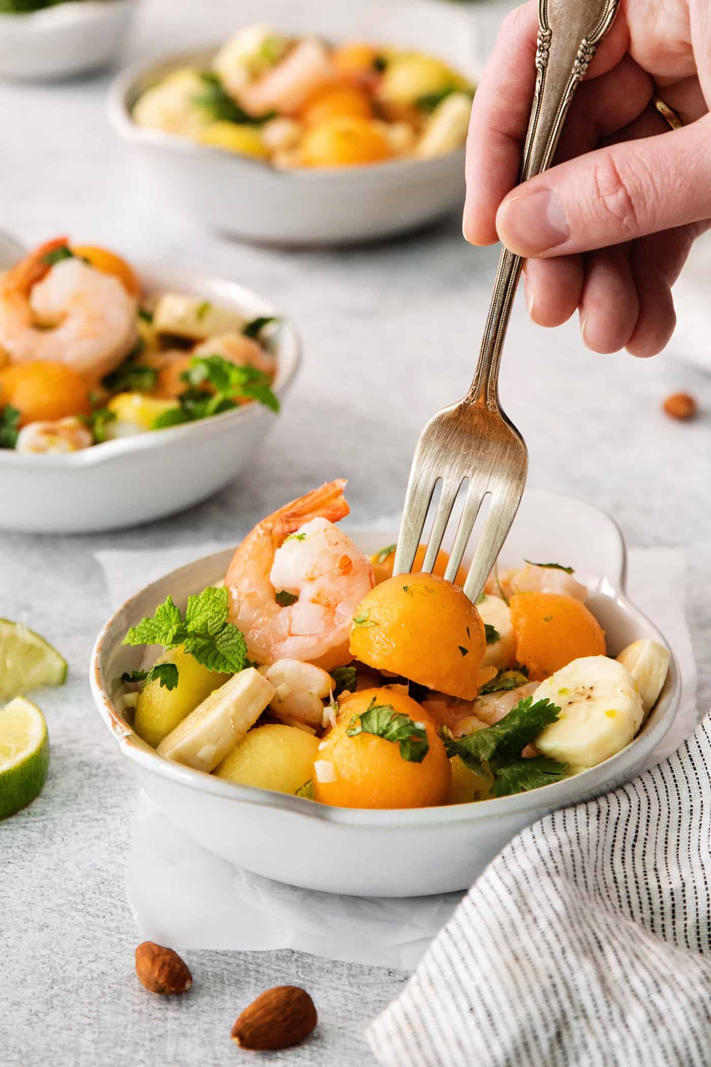 This Tropical Shrimp Salad has fresh, bright color and flavor. It's a unique, refreshing appetizer or salad recipe, great for entertaining! A hand holding a fork picks up a piece of melon out of a bowl of tropical shrimp salad with another bowl of salad shown in the background.