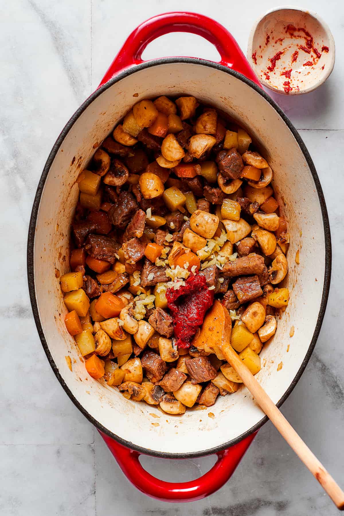 Comforting Beef Pot Pie is packed with tender beef and vegetables tucked into a stout-based gravy with flaky pie crust balanced on top. A wooden spoon stirs in tomato paste to a pot of beef pot pie filling.