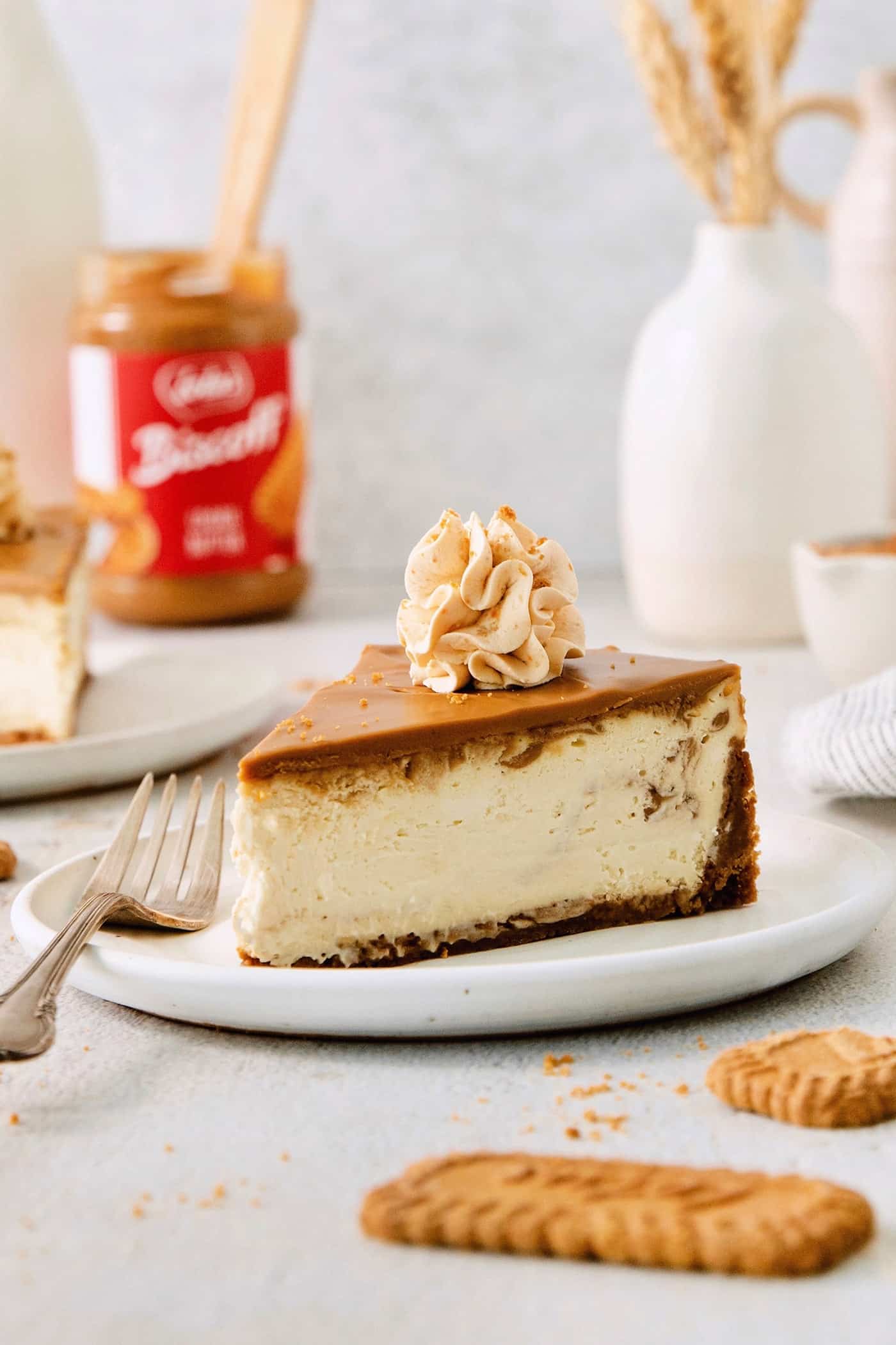 This Biscoff Cheesecake balances a rich cream cheese layer swirled with Biscoff cookie butter on a buttery cookie crust. A slice of biscoff cheesecake is shown on a plate with biscoff cookie butter in the background.