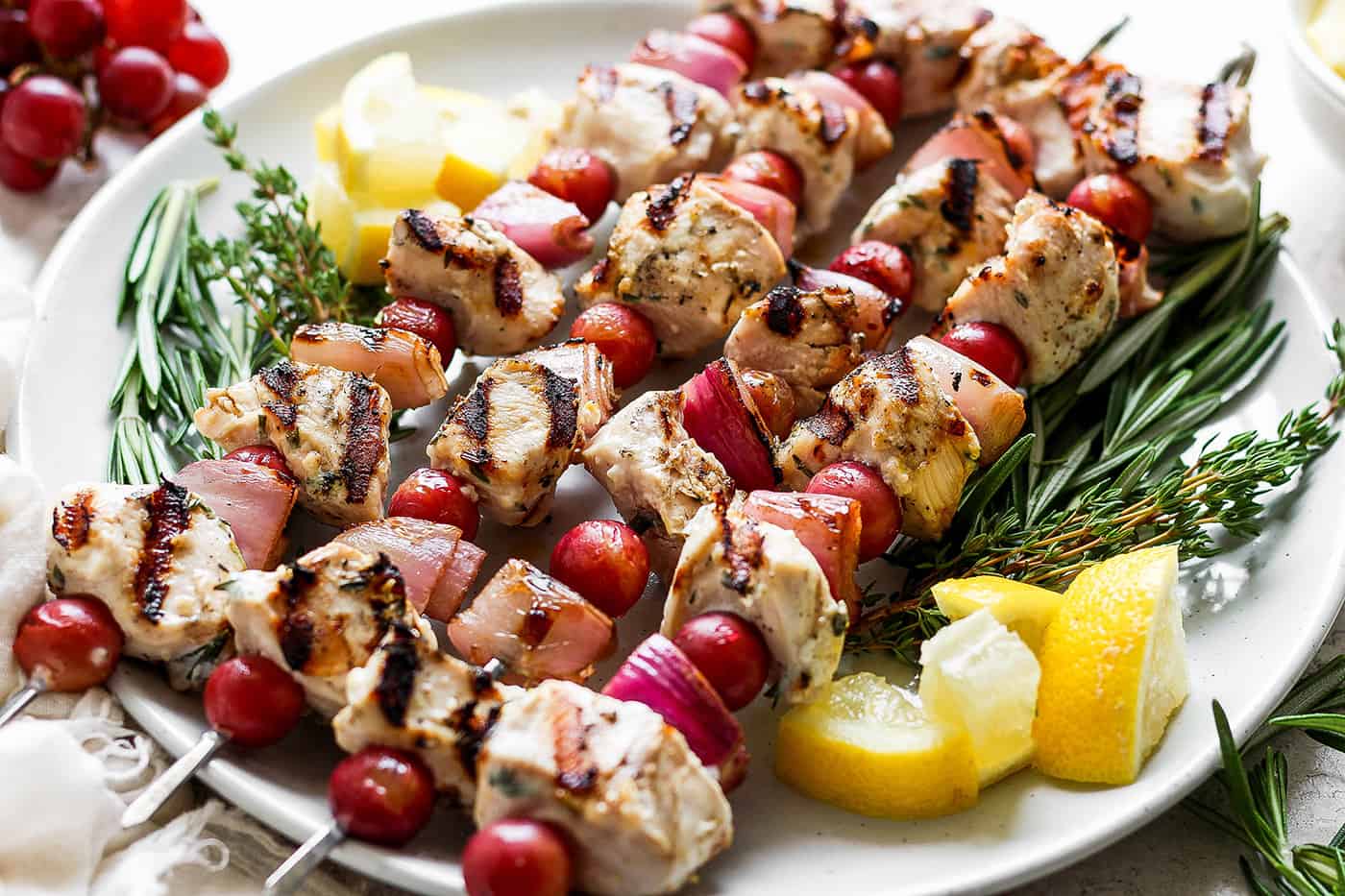 These tender Chicken Kabobs with Rapes are marinated in a lemon yogurt sauce and paired with honey balsamic shallots and sweet juicy grapes. A white plate holds chicken kabobs with grapes on skewers with lemon next to them.