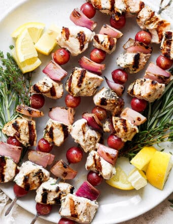 A white plate holds chicken kabobs with grapes on skewers with lemon next to them.