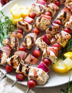 A white plate holds chicken kabobs with grapes on skewers with lemon next to them.