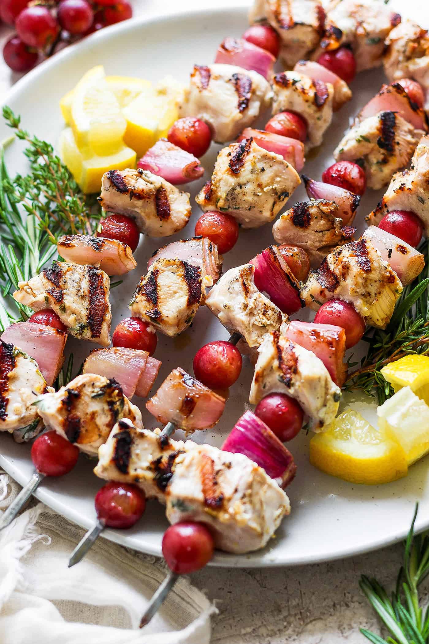 These tender Chicken Kabobs with Rapes are marinated in a lemon yogurt sauce and paired with honey balsamic shallots and sweet juicy grapes. A white plate holds chicken kabobs with grapes on skewers with lemon next to them.