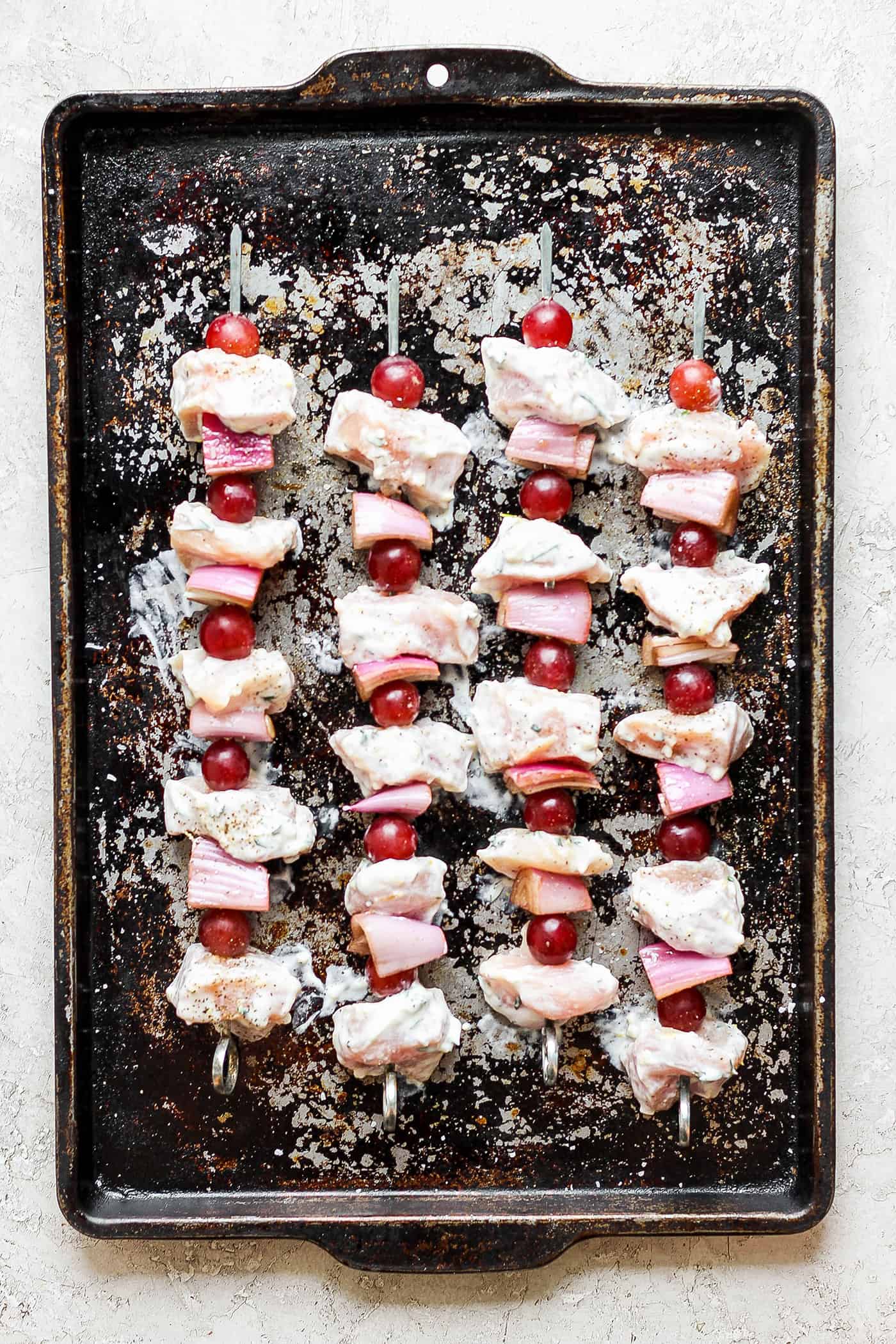 These tender Chicken Kabobs with Rapes are marinated in a lemon yogurt sauce and paired with honey balsamic shallots and sweet juicy grapes. A baking sheet holds chicken kabobs with grapes before they are cooked.