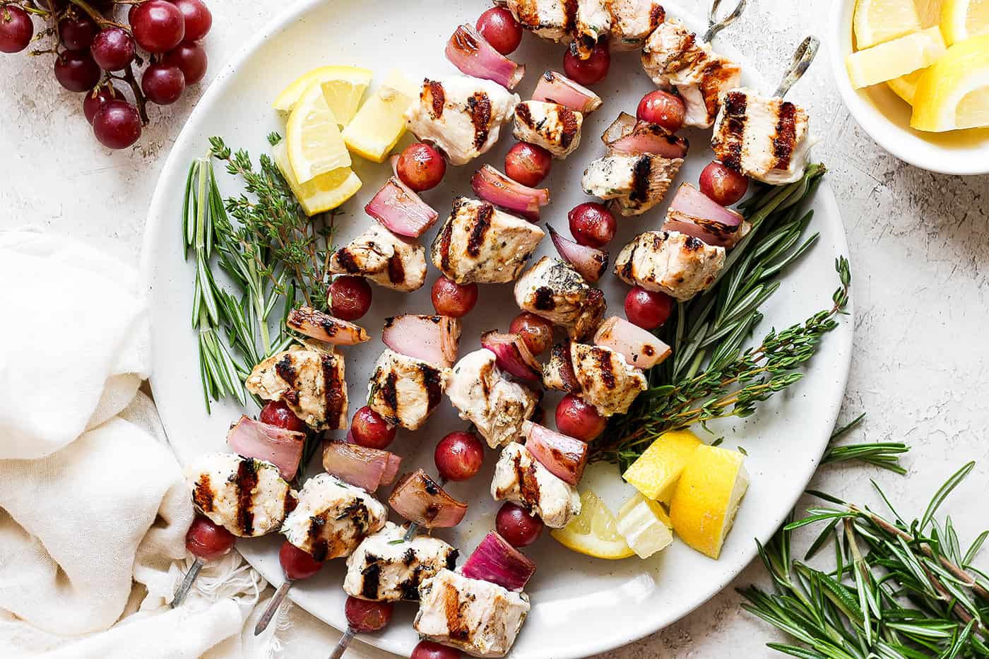 These tender Chicken Kabobs with Rapes are marinated in a lemon yogurt sauce and paired with honey balsamic shallots and sweet juicy grapes. A white plate holds chicken kabobs with grapes on skewers with lemon next to them.