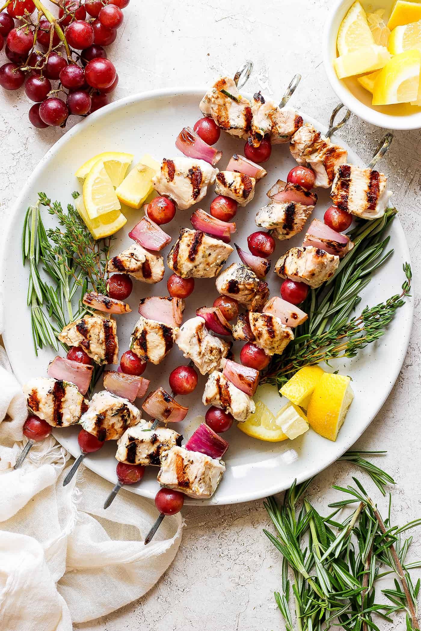 These tender Chicken Kabobs with Rapes are marinated in a lemon yogurt sauce and paired with honey balsamic shallots and sweet juicy grapes. A white plate holds chicken kabobs with grapes on skewers with lemon next to them.