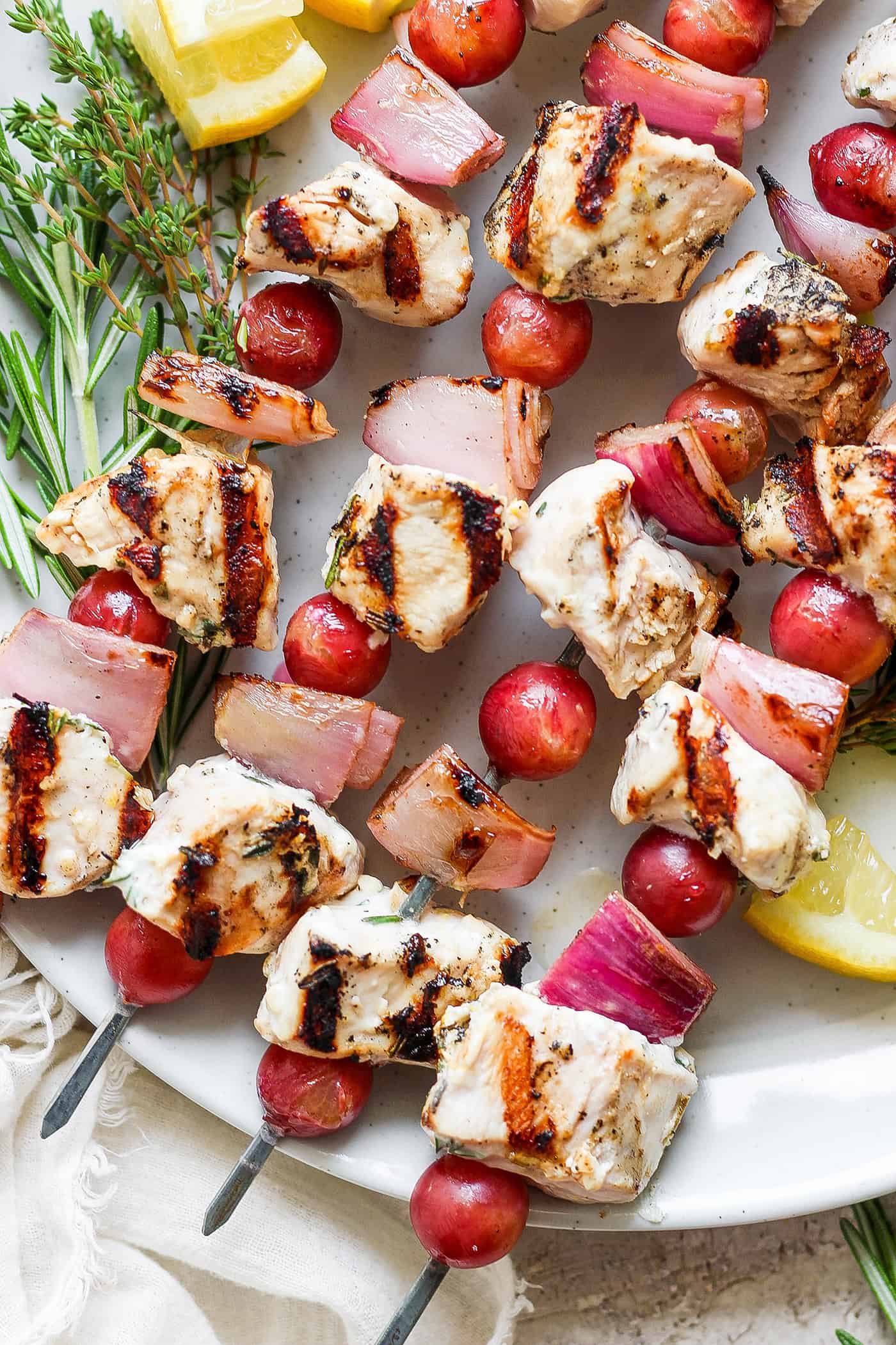 These tender Chicken Kabobs with Rapes are marinated in a lemon yogurt sauce and paired with honey balsamic shallots and sweet juicy grapes. A white plate holds chicken kabobs with grapes on skewers.