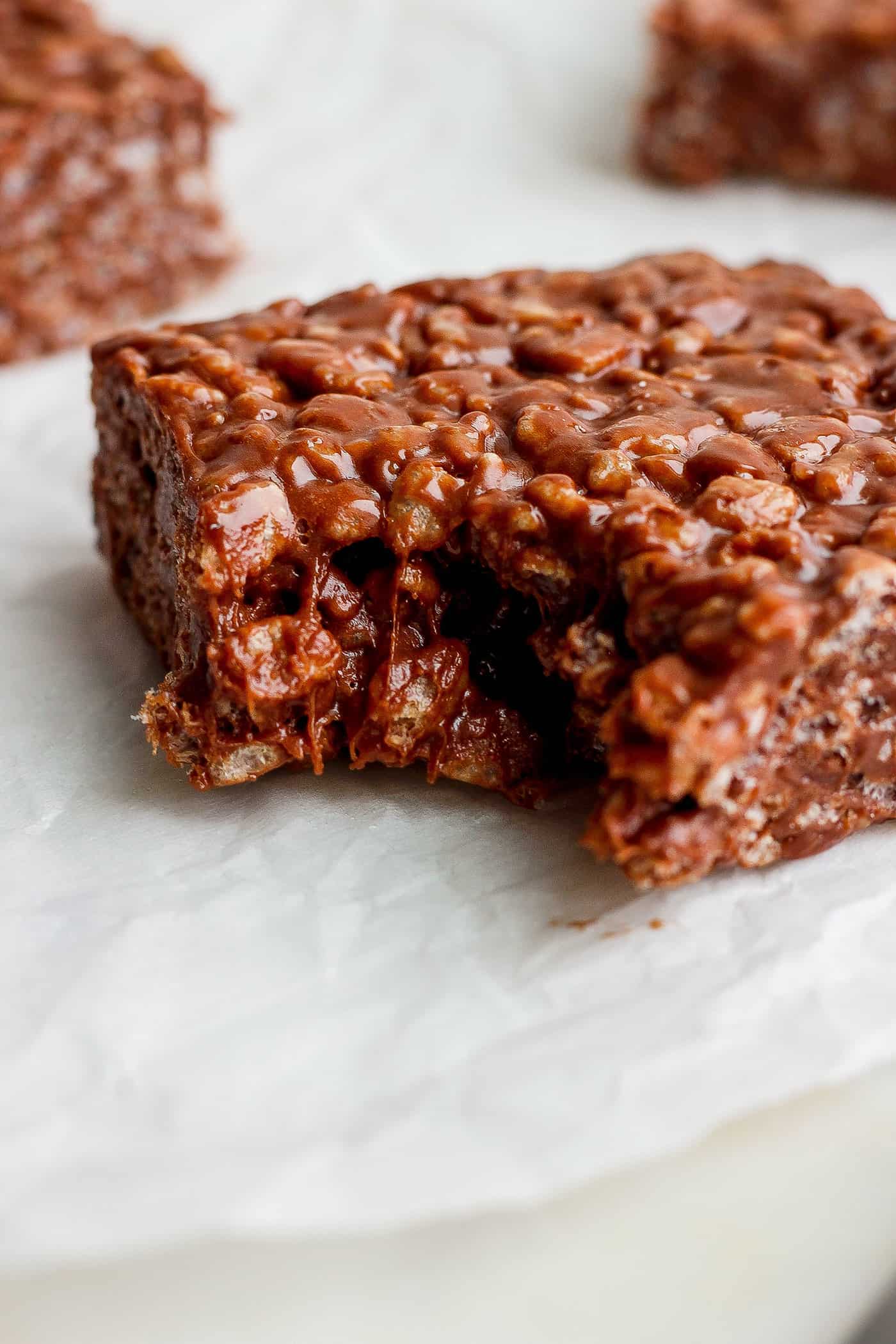 Chocolate Rice Krispie Treats have a chewy, fudge-like texture and are full of rich chocolate and sweet marshmallow flavor. A chocolate rice krispie treat is shown with a bite taken out of it on a white background.