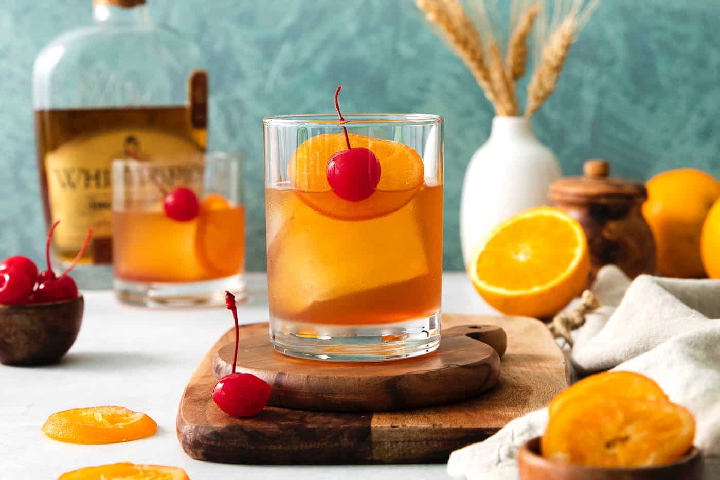 This classic Old Fashioned is a go-to before-dinner cocktail! It’s simple and easy, made with just three ingredients: whiskey, simple syrup, and bitters. An old fashioned drink is shown on a wood board with more drinks and oranges in the background.