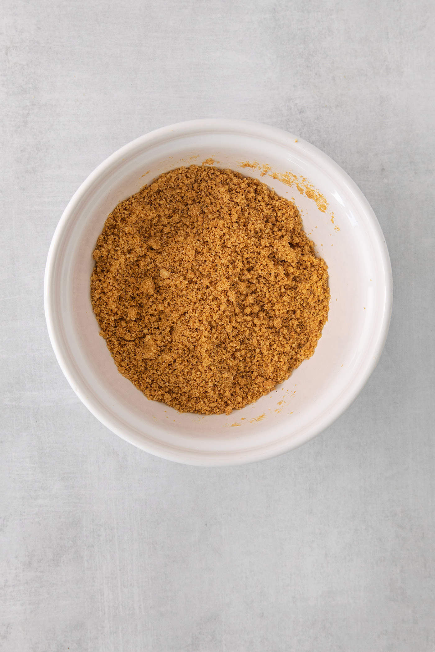 Crumbled graham crackers to make a crust for pumpkin cheesecake bars are shown in a white bowl.