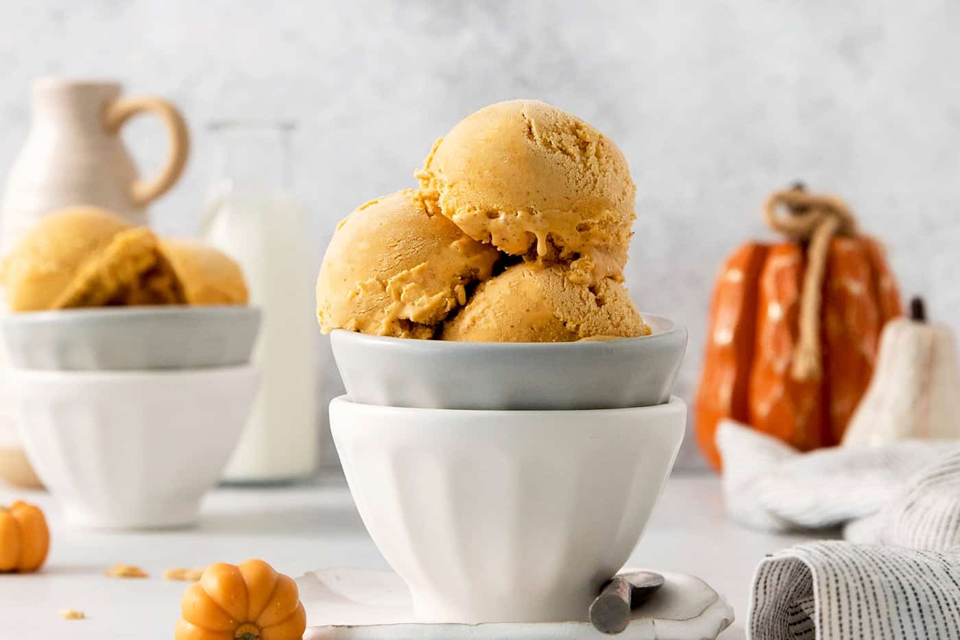 This Pumpkin Ice Cream is rich and creamy, flavored with pumpkin puree and pumpkin pie spice, and sweetened with brown sugar. Custard-based ice creams may seem intimidating, but this one is very easy and only requires a handful of ingredients. Three scoops of orange pumpkin ice cream are shown in a gray bowl stacked in a white bowl.