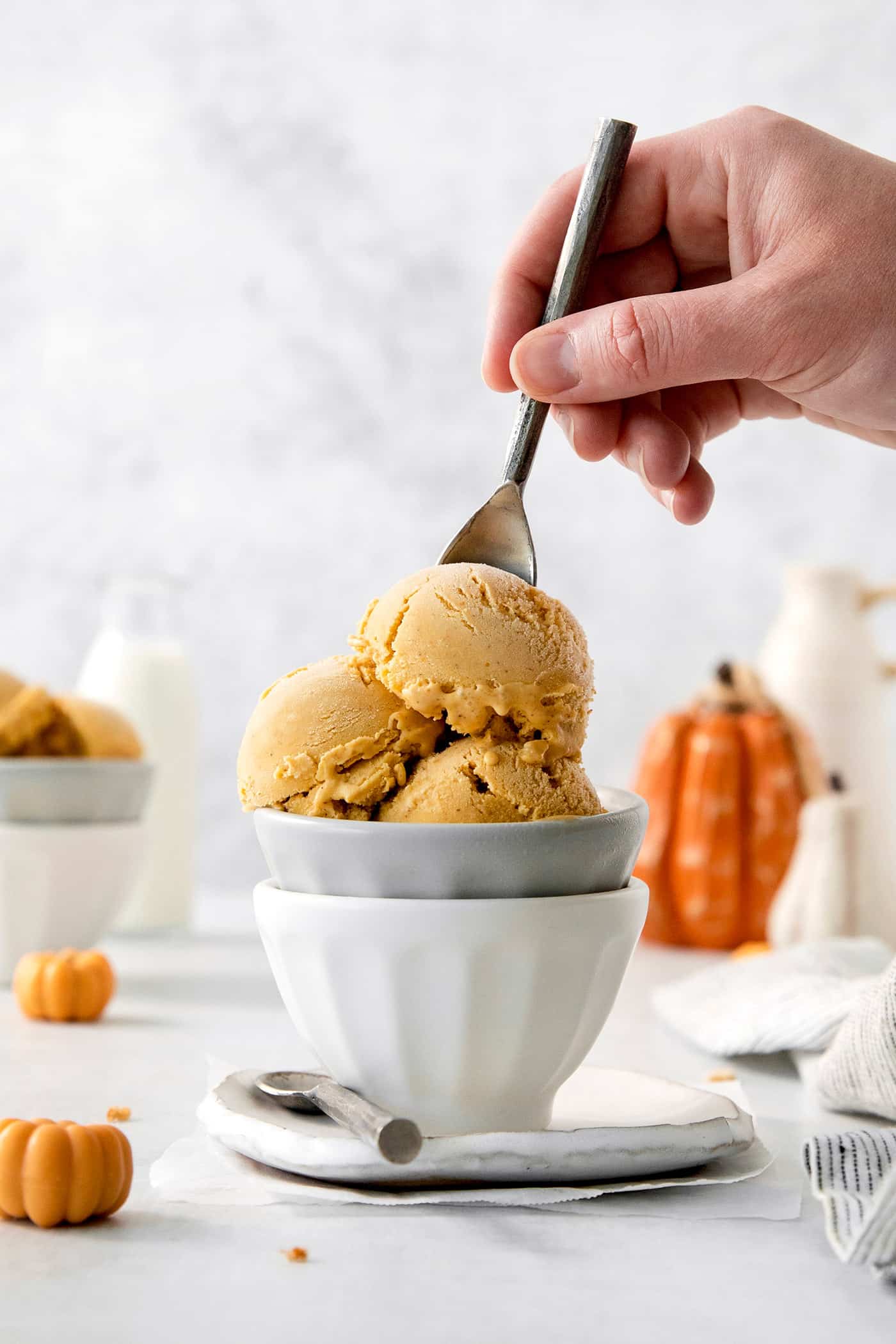 This Pumpkin Ice Cream is rich and creamy, flavored with pumpkin puree and pumpkin pie spice, and sweetened with brown sugar. Scoops of orange pumpkin ice cream are shown in a white and gray bowl as a hand dips in a spoon.