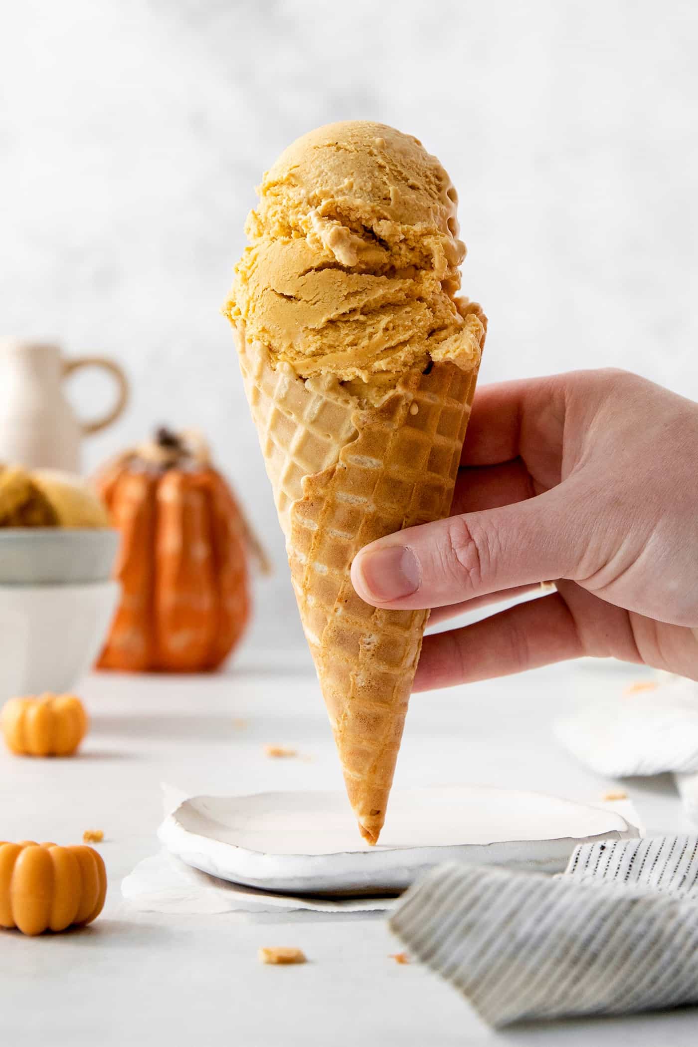 This Pumpkin Ice Cream is rich and creamy, flavored with pumpkin puree and pumpkin pie spice, and sweetened with brown sugar. Custard-based ice creams may seem intimidating, but this one is very easy and only requires a handful of ingredients. A hand holds a cone of pumpkin ice cream.