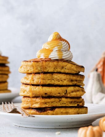 A stack of pumpkin pancakes topped with maple cinnamon whipped cream on a white plate.