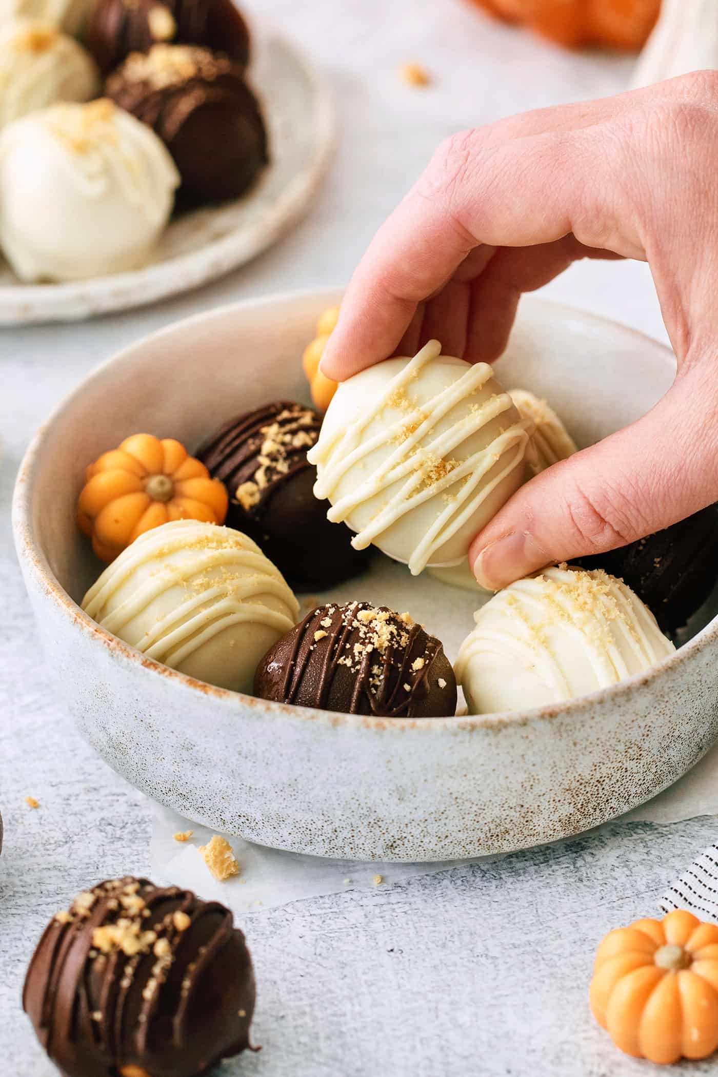 These Pumpkin Pie Truffles are the perfect fall treat! The truffle filling is made with crisp cookies, cream cheese, pumpkin puree, and spice. A hand picks up a white chocolate covered pumpkin pie truffle from a bowl of truffles.