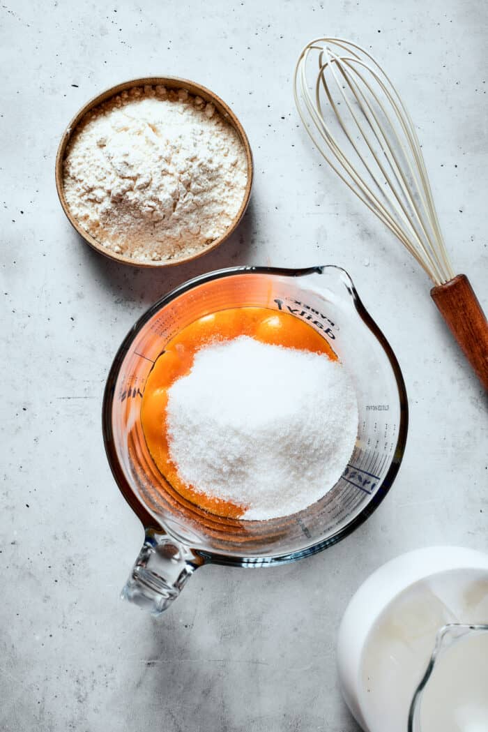 Napoleon Cake takes flaky homemade pastry and layers it with a sumptuous egg custard to make a creamy and cool dessert! Eggs and sugar are shown in a glass measuring cup with a whisk next to it.