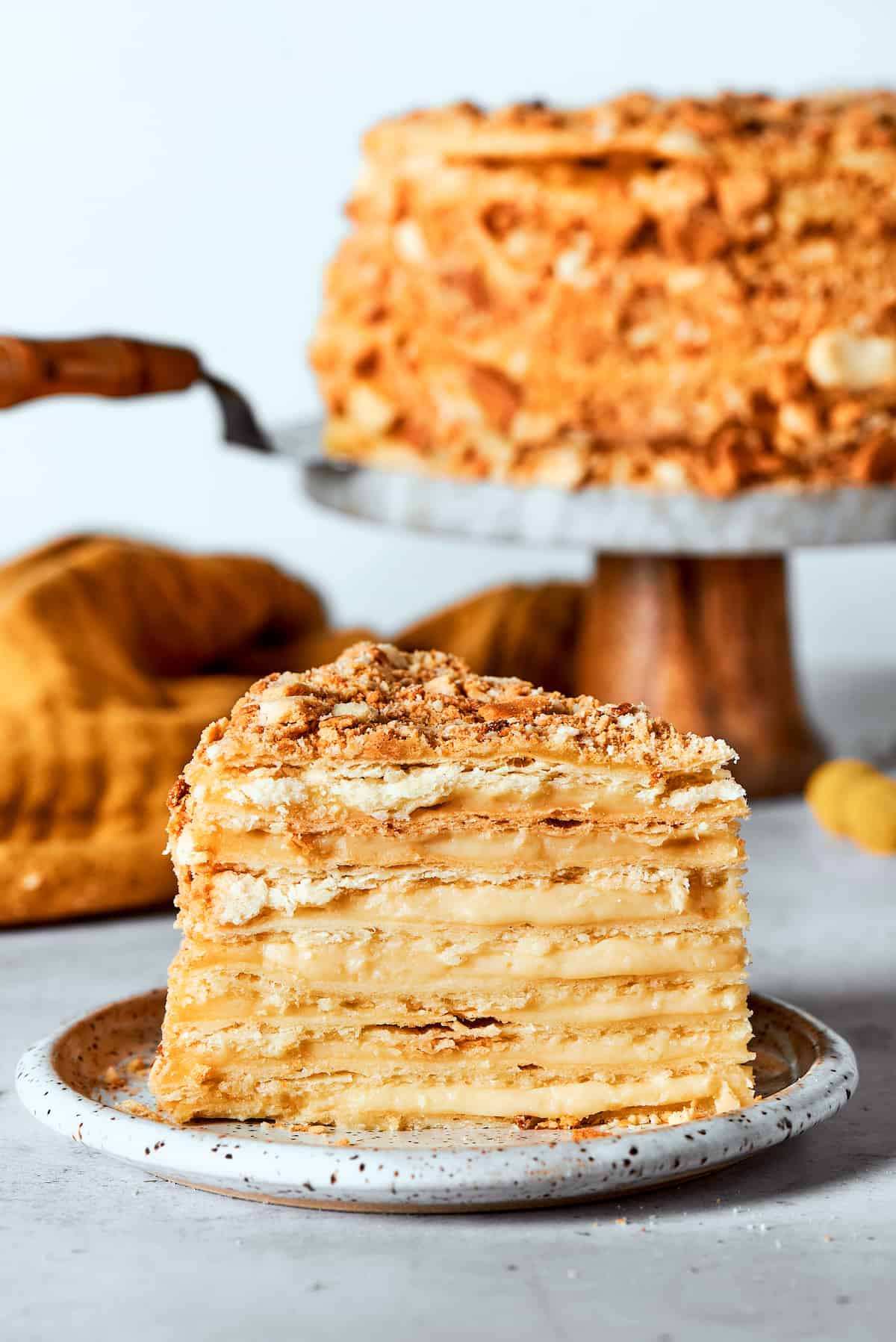 Napoleon Cake takes flaky homemade pastry and layers it with a sumptuous egg custard to make a creamy and cool dessert! A side view of a piece of Napoleon cake with the cake in the background.