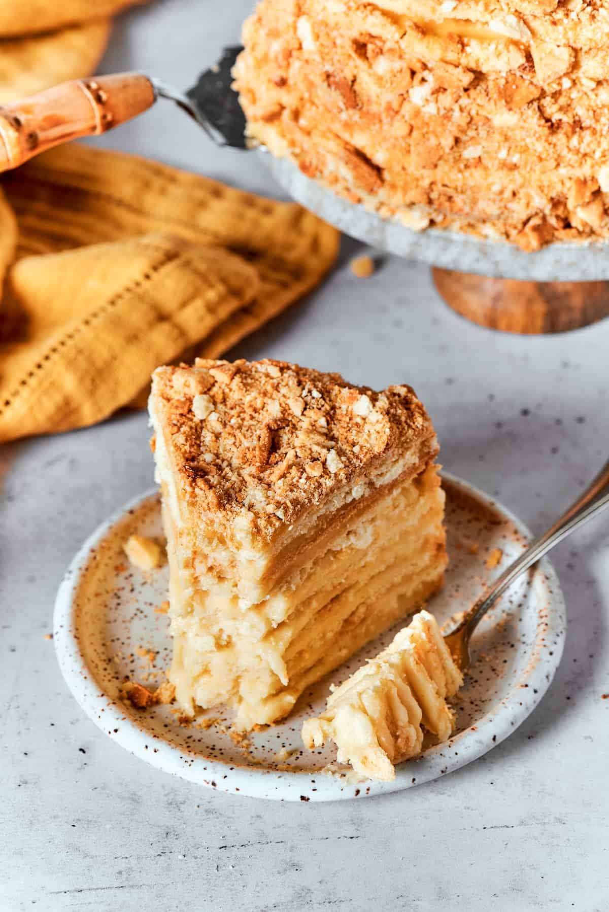 Napoleon Cake takes flaky homemade pastry and layers it with a sumptuous egg custard to make a creamy and cool dessert! A piece of Napoleon cake on a plate with a fork and an orange napkin next to it.