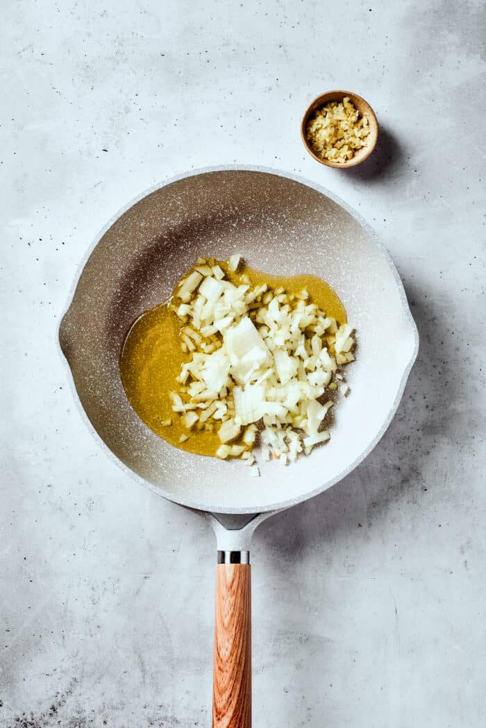 Easy, Italian-inspired Pasta Fazool is a wonderful soup that pairs creamy white beans with ground meat and pasta in a tomato-based broth. Onions and olive oil are shown in a white skillet.