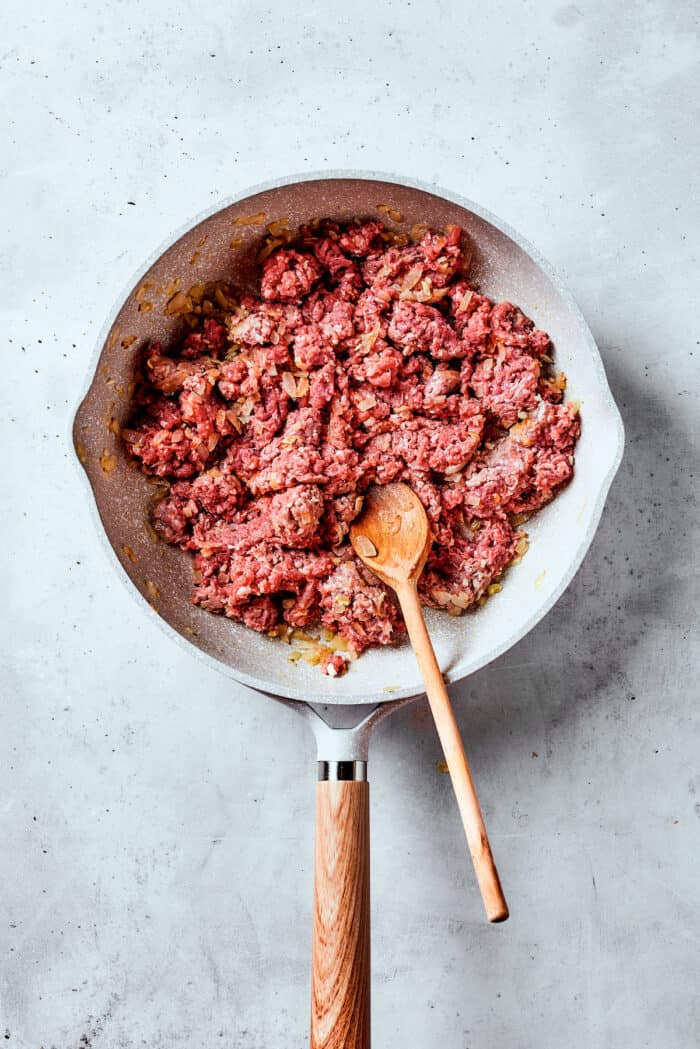 Easy, Italian-inspired Pasta Fazool is a wonderful soup that pairs creamy white beans with ground meat and pasta in a tomato-based broth. Meat cooks in a white skillet with a wooden spoon.