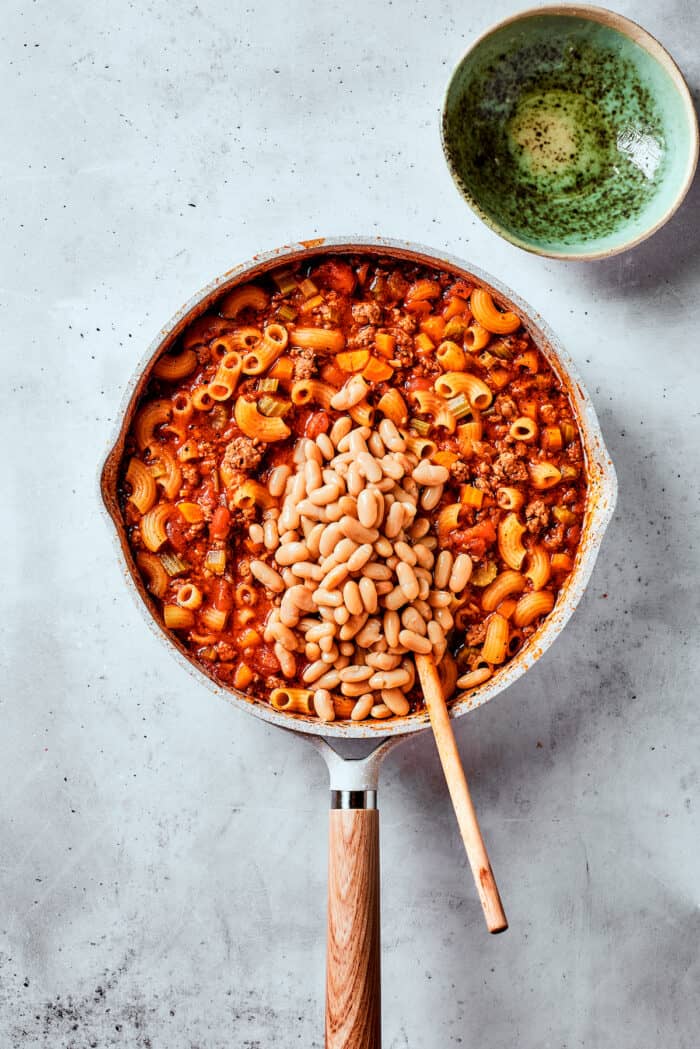 Easy, Italian-inspired Pasta Fazool is a wonderful soup that pairs creamy white beans with ground meat and pasta in a tomato-based broth. Beans are stirred into a pot of pasta fazool with a wooden spoon.