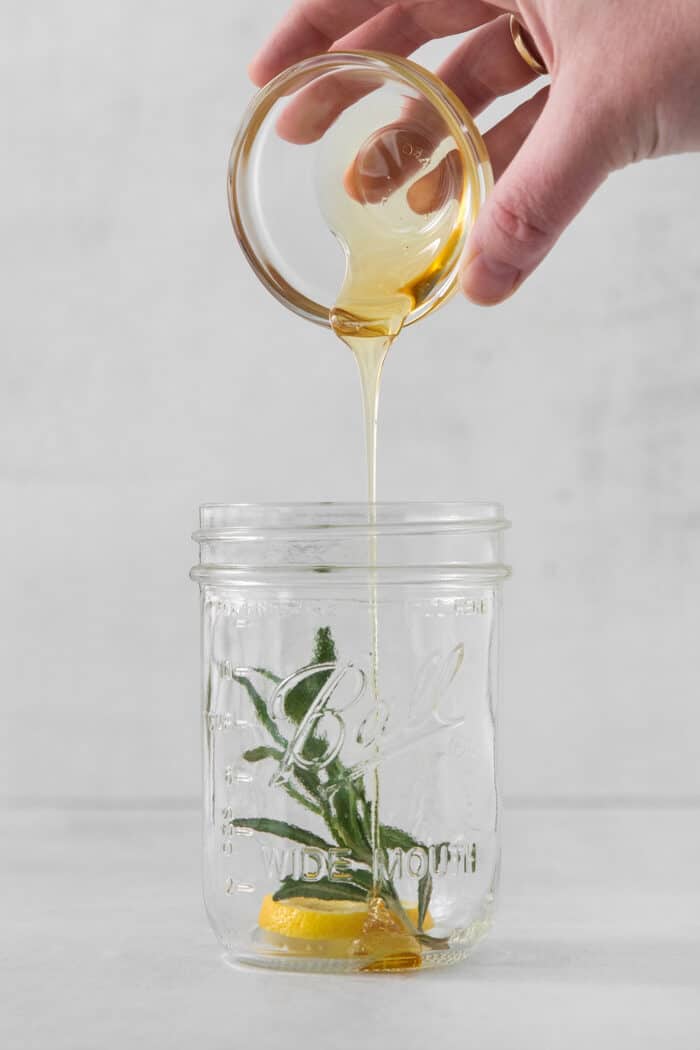 This Bourbon Limoncello Cocktail is a refreshing drink featuring fresh rosemary, lemon, honey, bourbon, and limoncello. A hand pours honey into a mason jar with rosemary in it.