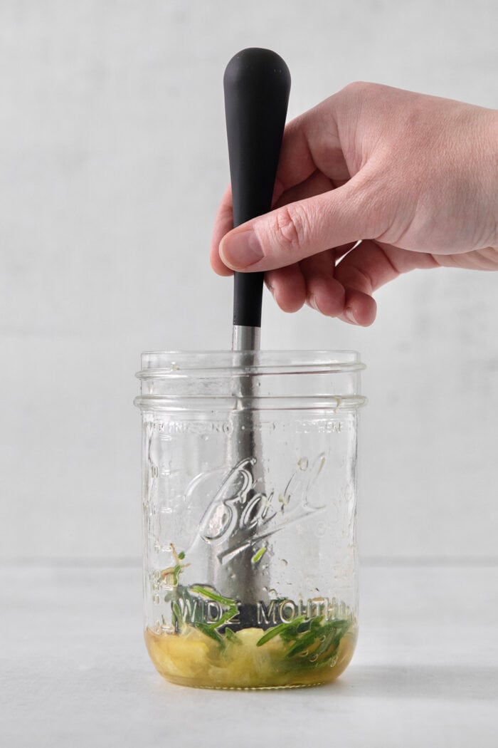 This Bourbon Limoncello Cocktail is a refreshing drink featuring fresh rosemary, lemon, honey, bourbon, and limoncello. A hand muddles rosemary, lemon, and honey together in a glass jar.
