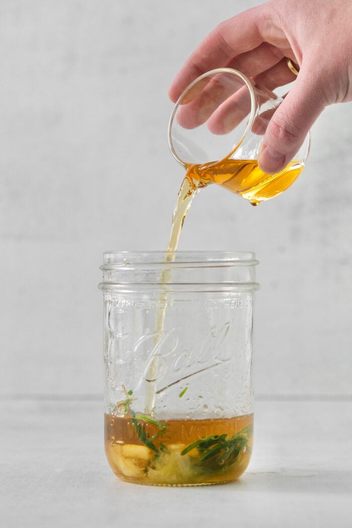 This Bourbon Limoncello Cocktail is a refreshing drink featuring fresh rosemary, lemon, honey, bourbon, and limoncello. A hand pours bourbon into a glass jar for bourbon limoncello cocktail.
