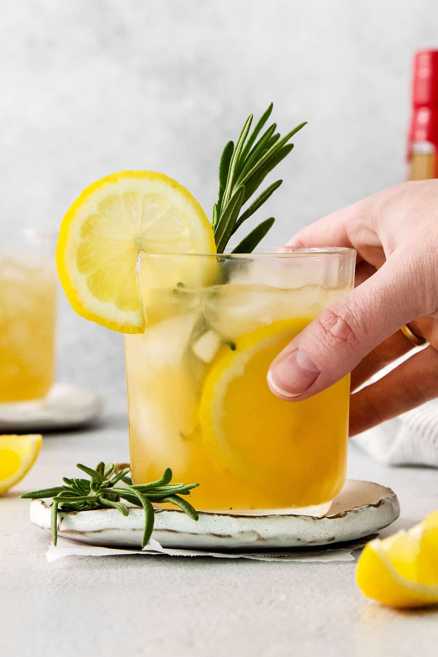 This Bourbon Limoncello Cocktail is a refreshing drink featuring fresh rosemary, lemon, honey, bourbon, and limoncello. A hand holds a glass of bourbon limoncello cocktail toped with a lemon wheel and fresh rosemary.
