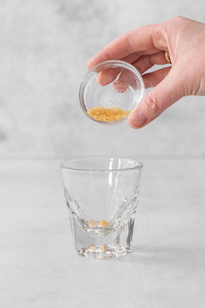 A Cortadito is a classic Cuban coffee made from equal parts sweetened espresso and frothed milk. It is rich, creamy, and sweet. A hand pours sugar into a small glass.