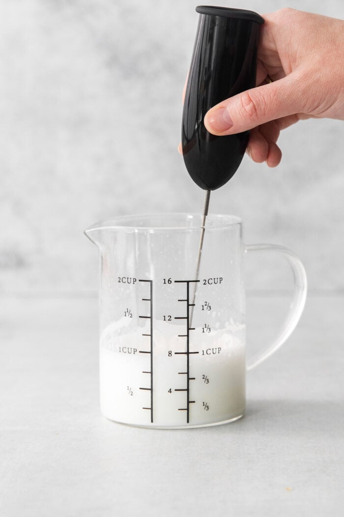 A Cortadito is a classic Cuban coffee made from equal parts sweetened espresso and frothed milk. It is rich, creamy, and sweet. A hand holds a milk frother to froth a pitcher of milk.
