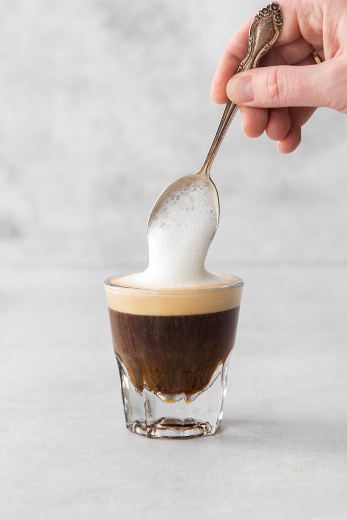 A Cortadito is a classic Cuban coffee made from equal parts sweetened espresso and frothed milk. It is rich, creamy, and sweet. A hand holding a spoon tops a cortadito with frothed milk.