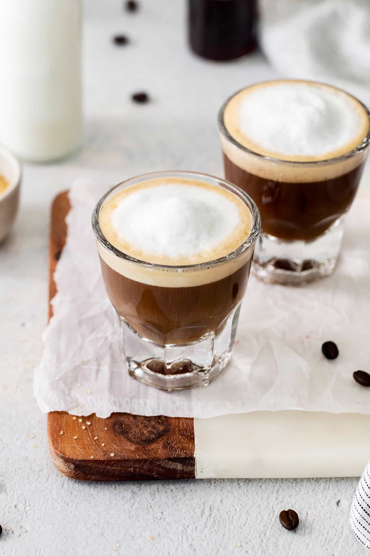 A Cortadito is a classic Cuban coffee made from equal parts sweetened espresso and frothed milk. It is rich, creamy, and sweet. Two cortaditos are shown on a board with milk in the background.